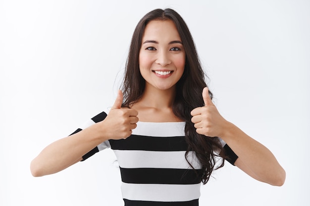 Photo gratuite fille brune asiatique confiante, motivée et solidaire en t-shirt rayé, montrant un geste du pouce levé pour encourager quelqu'un qu'il fait le mieux, accepter ou approuver quelque chose, comme une idée, un fond blanc