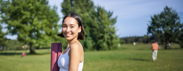 Photo gratuit une fille de fitness souriante avec un tapis en caoutchouc se tient dans le parc en uniforme pour l'entraînement et les activités sportives