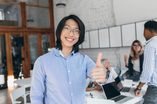 Photo gratuite garçon riant asiatique posant avec le pouce vers le haut au début de la journée de travail. employé de bureau chinois en chemise bleue et lunettes souriant avec ordinateur portable.