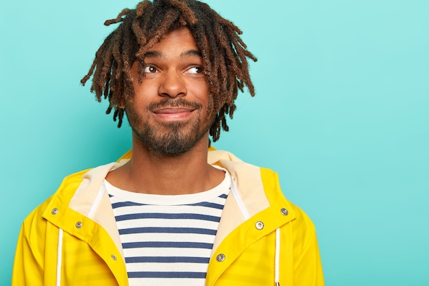 Photo gratuite un gars pensive attrayant avec un regard heureux, concentré sur le côté, a une moustache, une barbe et des redoutes, porte un pull et un imperméable de marin décontracté, pense à ses projets le week-end, se tient contre le mur bleu