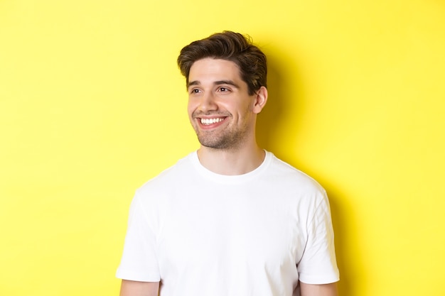 Photo gratuite gros plan d'un homme barbu attrayant en t-shirt blanc souriant, regardant à gauche à l'espace de copie, debout sur fond jaune.