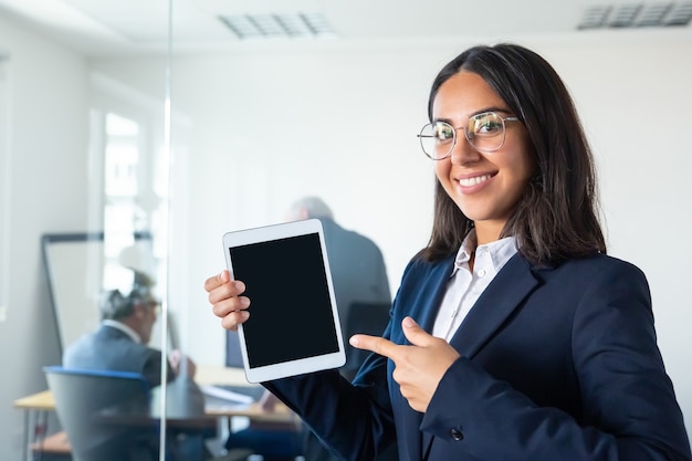 Heureuse femme d'affaires confiante tenant la tablette, montrant et pointant le doigt sur un écran vide, regardant la caméra et souriant. Copiez l'espace. Concept de communication et de publicité