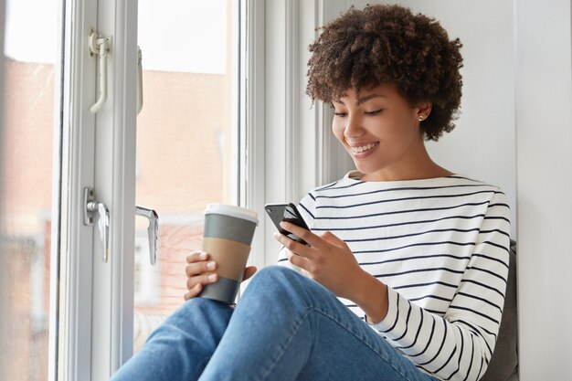 Heureuse femme à la peau sombre avec une expression heureuse, lit un livre en ligne via un téléphone portable, vérifie le courrier électronique dans les réseaux sociaux