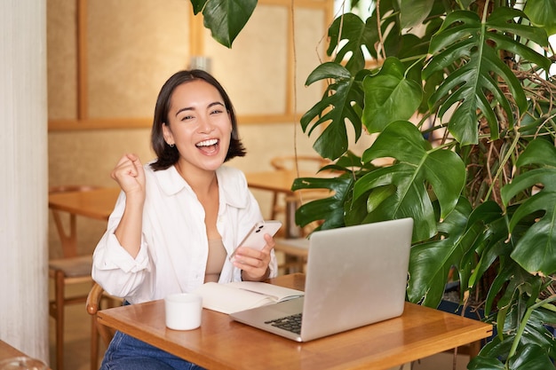 Photo gratuite heureuse jeune femme asiatique se sent gagnante et célébrant le succès assis avec un ordinateur portable et un smartphone en c