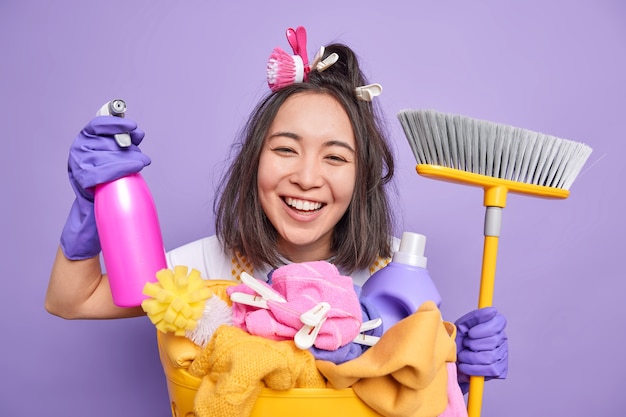 Photo gratuite heureuse jeune femme au foyer asiatique sourit largement aide à propos de la maison porte des gants en caoutchouc contient un détergent de nettoyage et un balai pour balayer les poses de sol près du panier à linge isolé sur fond violet