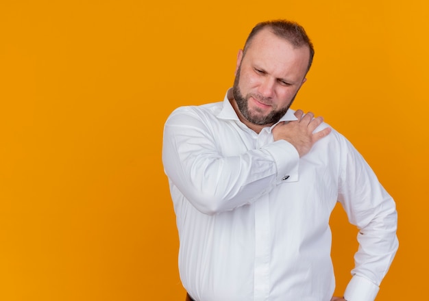 Photo gratuite homme barbu portant une chemise blanche à mal en touchant son épaule ressentant de la douleur debout sur un mur orange