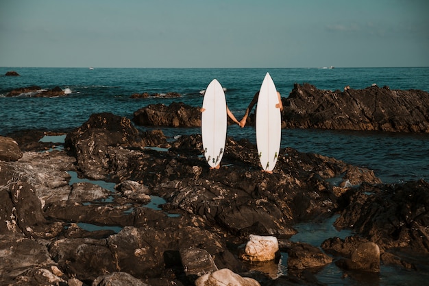 Photo gratuite homme et femme avec des planches de surf, main dans la main sur la côte de pierre