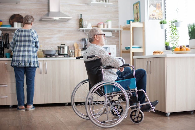 Homme handicapé assis en fauteuil roulant dans la cuisine regardant par la fenêtre pendant que sa femme prépare le petit-déjeuner. Invalide, retraité, handicapé, paralysie.