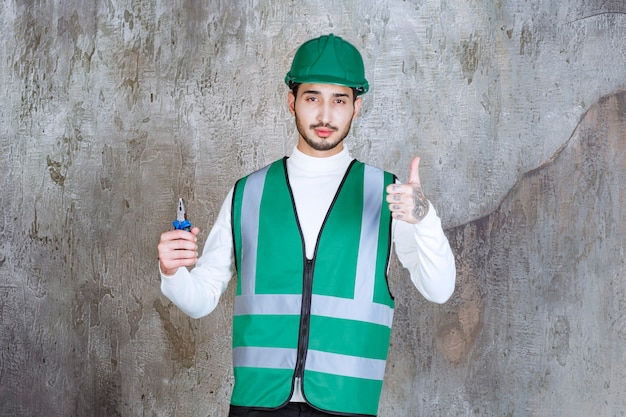 Photo gratuite homme ingénieur en uniforme jaune et casque tenant une pince bleue pour réparation et montrant un signe de main positif.