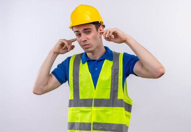 Photo gratuite homme jeune constructeur portant l'uniforme de construction et un casque de sécurité couvrant ses oreilles avec les doigts