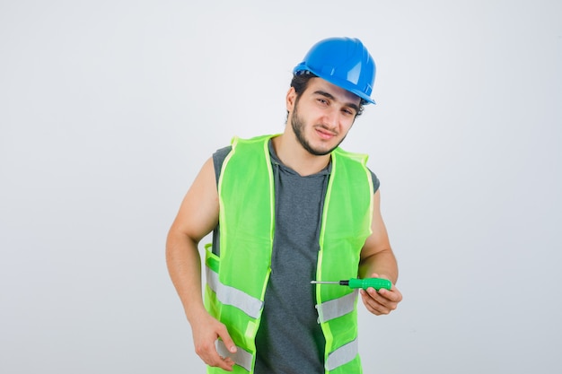 Photo gratuite homme jeune constructeur tenant un tournevis tout en regardant ailleurs en uniforme et à la vue de face, confiant.