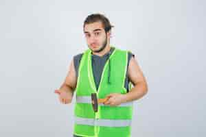 Photo gratuite homme jeune constructeur en uniforme de vêtements de travail tenant un marteau tout en montrant quelque chose et à la vue indécise, de face.