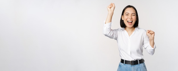 Photo gratuit image d'une heureuse femme asiatique chanceuse hourra geste gagnant et célébrant triomphant élevant hadns et riant debout sur fond blanc