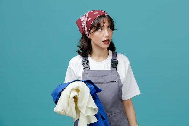 Photo gratuite impressionné jeune femme nettoyeuse portant un bandana uniforme et des gants en caoutchouc tenant des vêtements sales regardant le côté isolé sur fond bleu