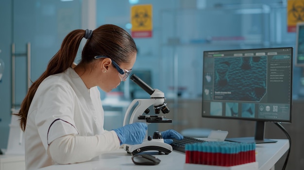 Photo gratuite ingénieur scientifique utilisant un microscope pour analyser un échantillon d'adn en laboratoire. femme biologiste travaillant avec une lentille en verre microscopique sur un outil de laboratoire et un ordinateur, portant des lunettes et des gants de protection.