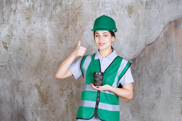 Photo gratuite ingénieure en uniforme vert et casque tenant une tasse de café noir et appréciant le produit