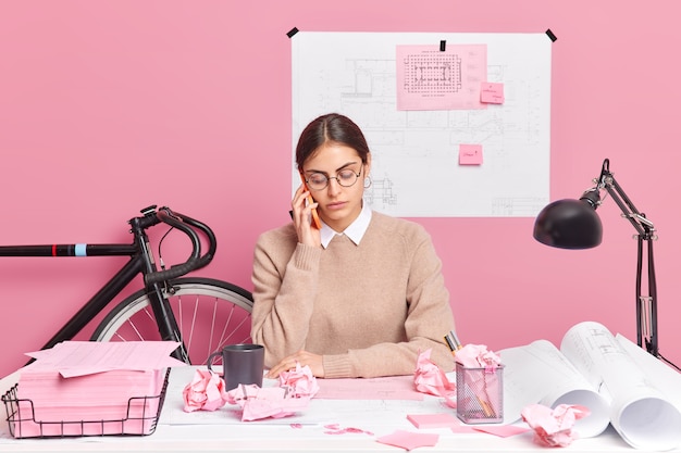 Photo gratuit une jeune architecte talentueuse fait des croquis de la planification du bâtiment a une conversation téléphonique impliquée dans le processus de travail entourée de papiers plans poses sur le bureau