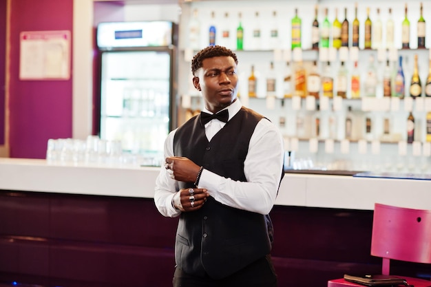 Photo gratuit jeune bel homme africain vêtu d'une chemise blanche, d'un gilet noir et d'un nœud papillon posés contre le comptoir du bar à la discothèque