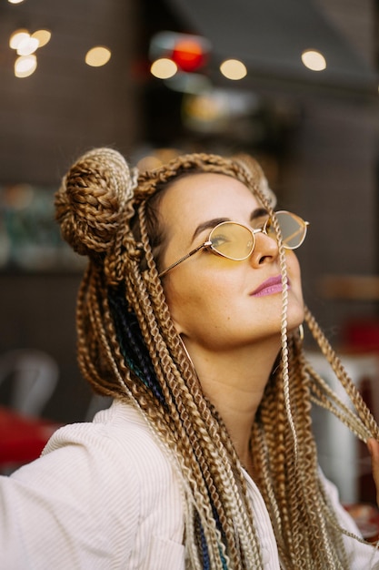 Photo gratuit jeune belle femme multiraciale avec une coiffure afro blonde tresses zizi sur le café de la rue. lunettes de soleil jaunes, maquillage lumineux, style hippie