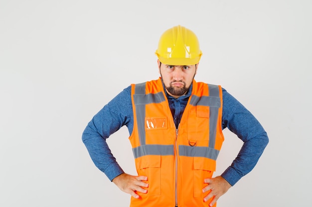 Photo gratuite jeune constructeur debout avec les mains sur la taille en chemise, gilet, casque et à la vue de face, nerveux.