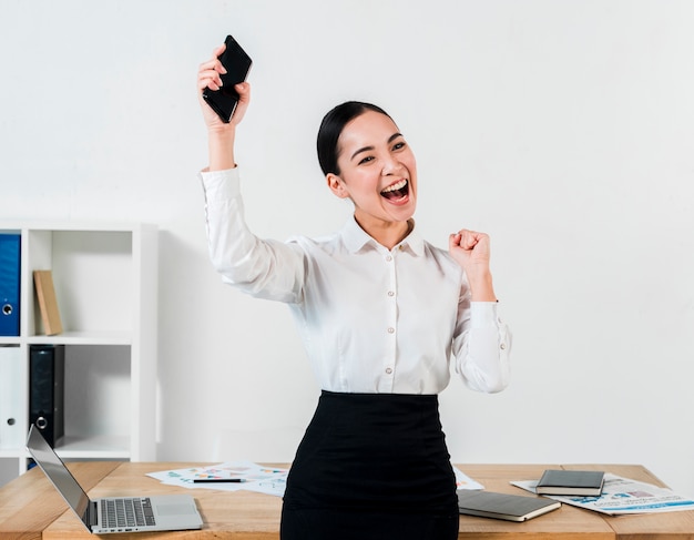 Photo gratuite jeune femme d'affaires réussie tenant mobile dans la main, serrant son poing au lieu de travail