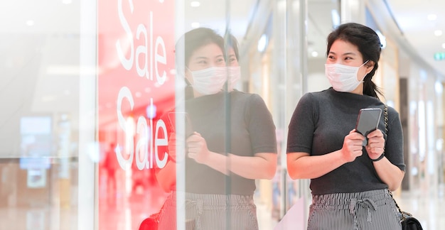 Photo gratuite jeune femme asiatique porter un masque facial protection main tenir smartphone debout regarder la signalisation du magasin montrer la promotion de la vente nouveau mode de vie d'achat normal après la propagation de covid19