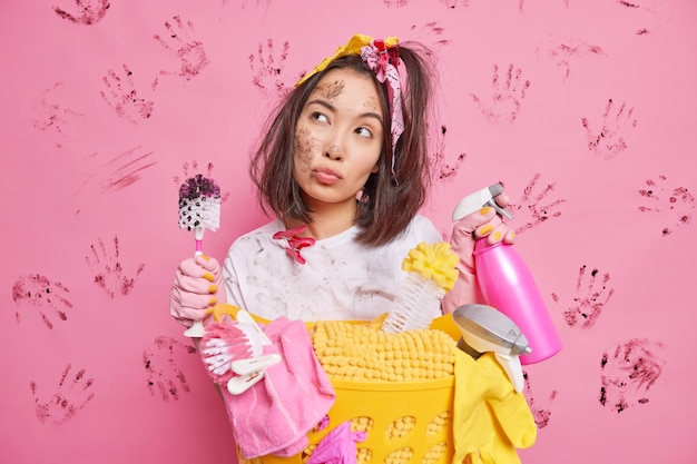 Photo gratuite une jeune femme au foyer asiatique sérieuse et réfléchie tient un détergent et une brosse a l'air pensif a des poses de visage sale près d'un panier de linge isolé sur un mur rose