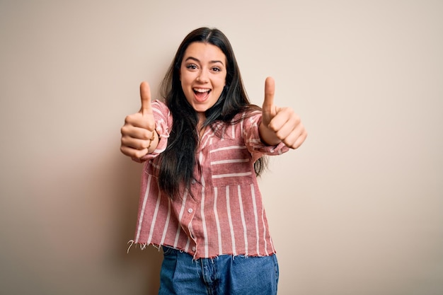 Photo gratuite jeune femme brune portant une chemise rayée décontractée sur fond isolé approuvant de faire un geste positif avec la main pouce levé souriant et heureux pour le succès geste du gagnant