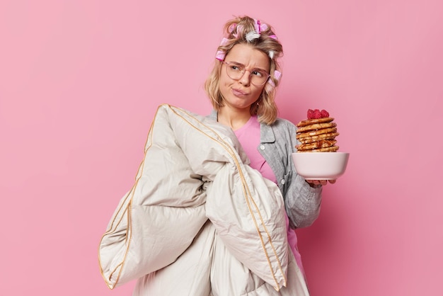 Une jeune femme insatisfaite tient une couette et des crêpes regarde malheureusement loin des sacs à main les lèvres ont une expression de mécontentement se réveille de mauvaise humeur porte un pyjama rend la coiffure bouclée subit des soins de beauté