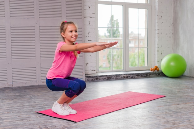 Jeune fille souriante accroupie et faisant de l&#39;exercice sur un tapis rose