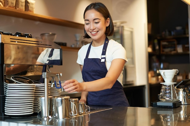 Photo gratuit jeune fille souriante barista dans un café préparant un cappuccino dans une machine à café du lait fumant portant un uniforme ap