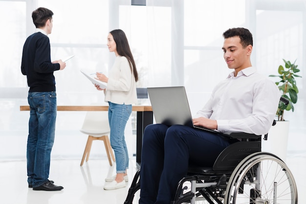 Photo gratuite jeune homme d'affaires assis sur un fauteuil roulant à l'aide d'un ordinateur portable avec son collègue discutant de quelque chose à l'arrière-plan