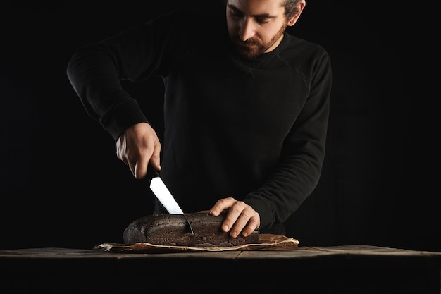 Photo gratuite jeune homme barbu en sueur noir utilise un grand couteau en chef pour trancher du pain de luxe fait maison à partir de figues et de seigle en papier kraft sur une table en bois rustique isolée sur fond noir