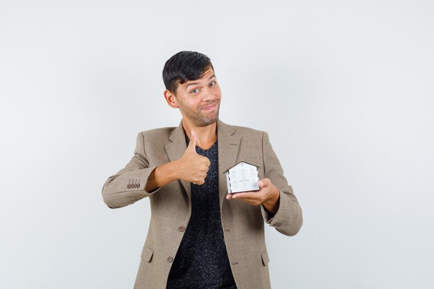 Photo gratuit jeune homme tenant un modèle de maison avec le pouce vers le haut en veste, t-shirt et à la joyeuse vue de face