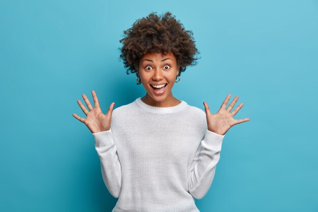 Jolie fille amusée aux cheveux afro bouclés, lève les paumes, a une expression joyeuse, sourit largement, voit quelque chose de drôle, porte un pull blanc,