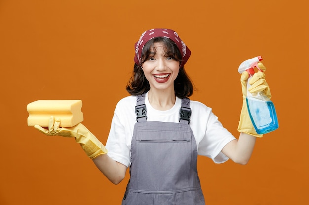 Photo gratuite joyeuse jeune femme nettoyante portant des gants en caoutchouc uniformes et un bandana tenant une éponge et un nettoyant regardant la caméra isolée sur fond orange