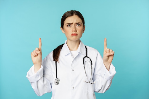 Photo gratuit médecin triste et sombre, travailleuse médicale boudant, pointant du doigt et levant les yeux, debout sur fond de torquoise