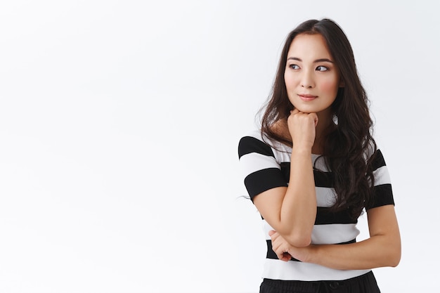 Photo gratuite pensive, séduisante et réfléchie, jeune femme brune asiatique en t-shirt rayé, tenir la main sous le menton, détournant le regard rêveuse, souriante en contemplant quelqu'un qui passe, debout sur fond blanc