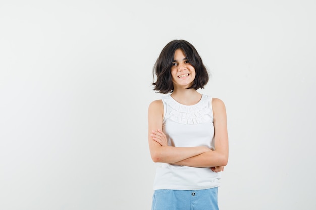 Photo gratuite petite fille debout avec les bras croisés en chemisier blanc, short et à la joyeuse. vue de face.