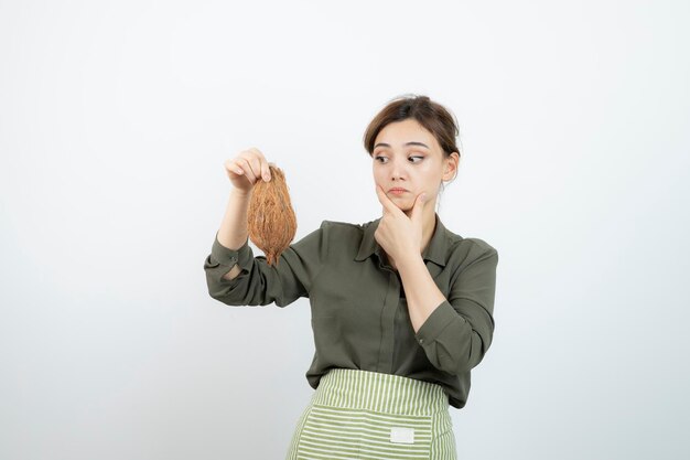 Photo de jeune femme en tablier tenant une noix de coco contre un mur blanc. Photo de haute qualité