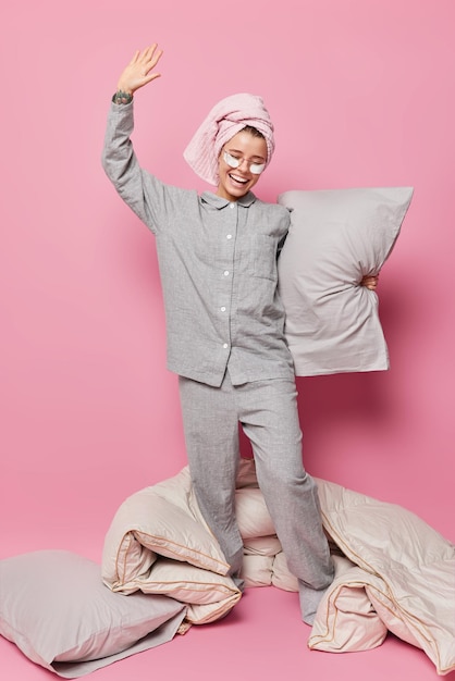 Photo gratuite la photo verticale d'une jeune femme heureuse s'amuse à se réveiller après un long sommeil se sent paisible et sereine porte un masque de sommeil et des vêtements de nuit tiennent des oreillers doux sur fond rose plein d'énergie