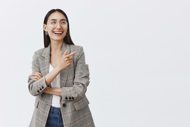 Photo gratuit plan horizontal d'une femme féminine élégante à lunettes et boucles d'oreilles à la mode, riant de plaisir et de bonheur tout en regardant et en pointant vers le coin supérieur droit, s'amusant sur un mur gris