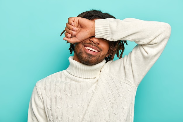 Photo gratuite plan horizontal d'un homme afro heureux avec des dreadlocks, couvre les yeux, cache le visage avec le bras, sourit joyeusement, porte un pull blanc, pose sur fond bleu.