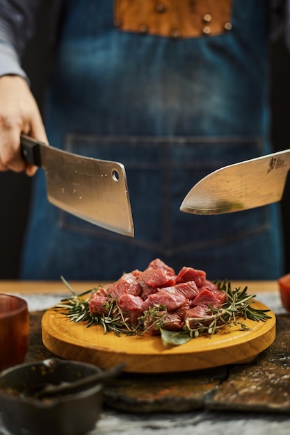 Photo gratuite plan vertical de deux couteaux dans les mains du cuisinier au-dessus de la viande crue coupée sur une assiette en bois