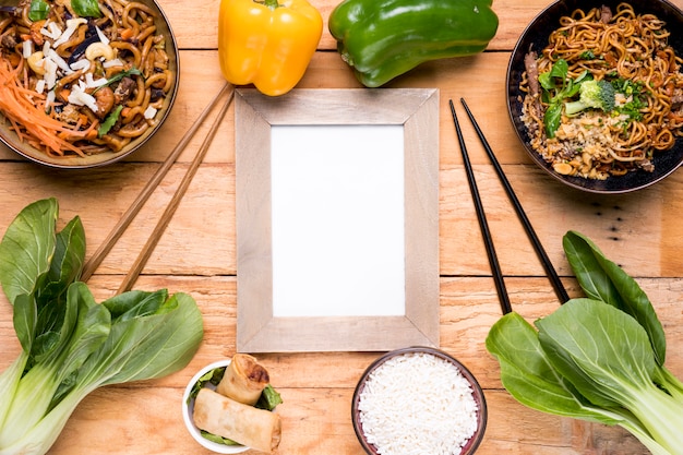 Photo gratuite poivrons; bokchoy; un bâton de coupe; rouleaux de printemps; bol de nouilles de riz et udon sur le bureau en bois