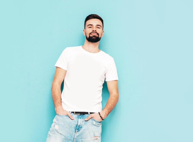 Portrait de beau modèle confiant hipster élégant hipster lambersexuel Homme vêtu d'un t-shirt blanc et d'un jean Mode homme isolé sur mur bleu en studio