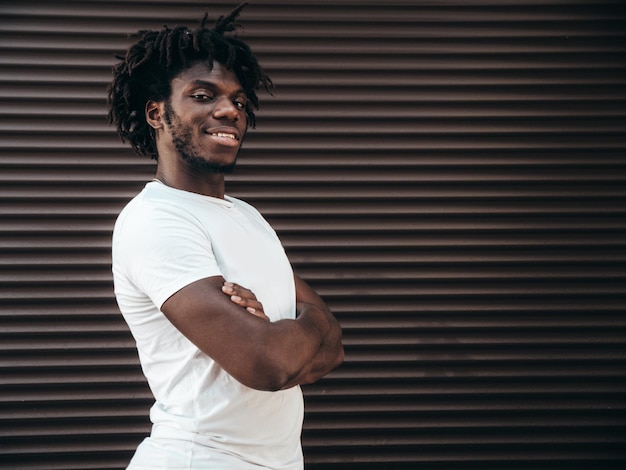 Photo gratuite portrait d'un beau modèle hipster souriant un homme africain mal rasé vêtu d'un t-shirt d'été blanc et d'un jean mode masculine avec une coiffure de dreadlocks posant sur le fond de la rue