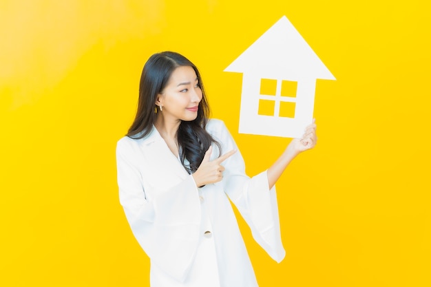 Photo gratuit portrait d'une belle jeune femme asiatique avec une maison en papier sur un mur jaune