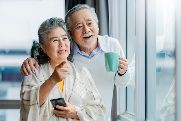 Photo gratuite portrait d'un couple de retraités senior asiatique souriant et regardant par la fenêtre de l'appartement tout en tenant l'épaule de sa femme