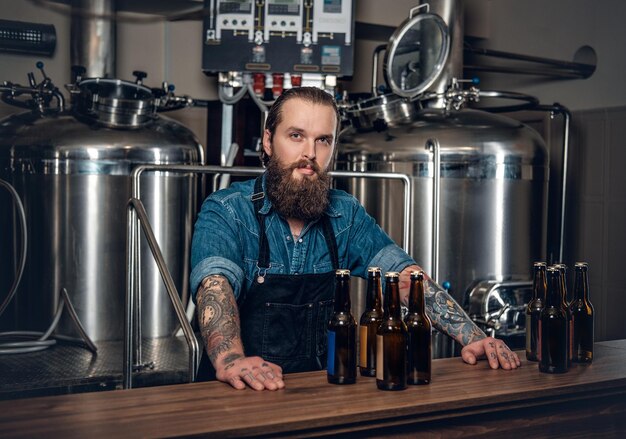 Portrait d'un fabricant masculin hipster tatoué et barbu présentant de la bière dans la microbrasserie.